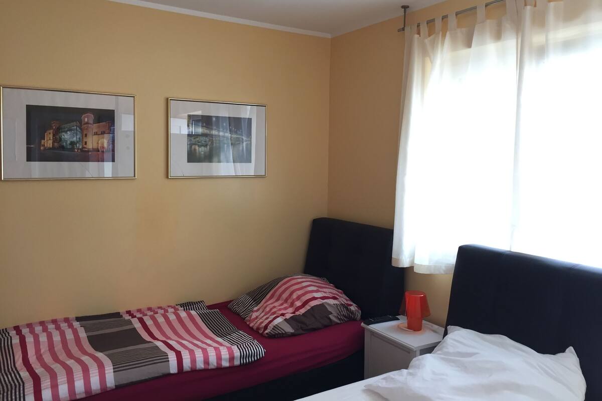 A single bed with a red and white striped blanket, a black headboard, and a white pillow. There is a small bedside table with a lamp and a remote control. Two framed pictures are hung on the wall above the bed.