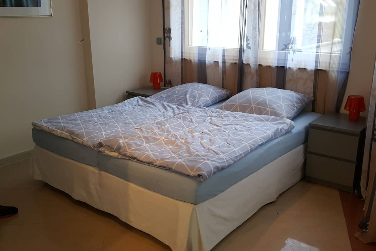 A bed with a geometric patterned bedspread, a nightstand with a red lamp, and a window with floral curtains.