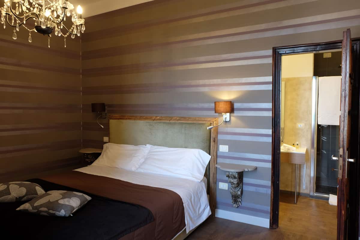 Una habitación de hotel con cama, candelabro, mesita de noche y baño visible a través de una puerta abierta.