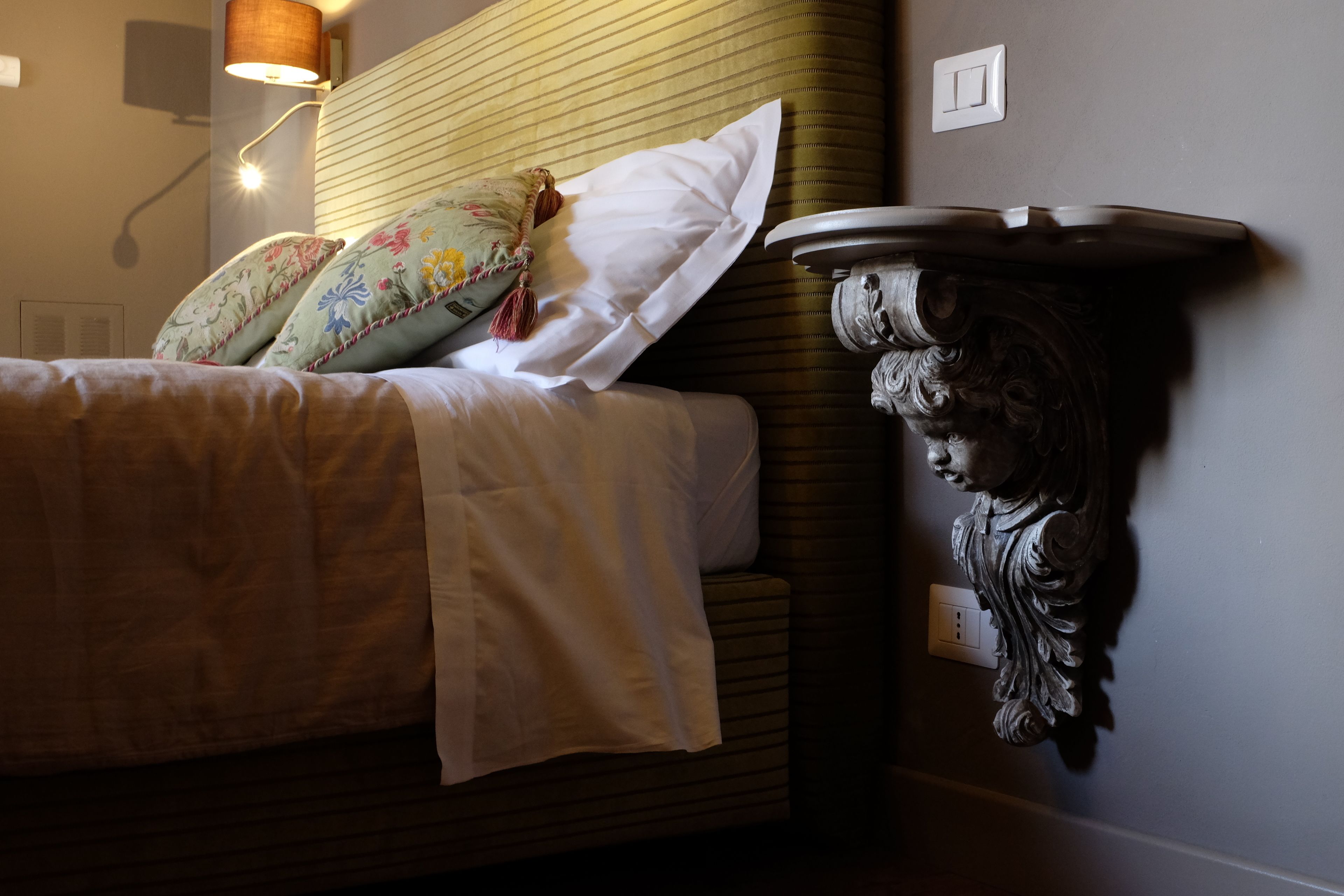A neatly made bed with floral pillows, a wall-mounted lamp, and a decorative wall piece.