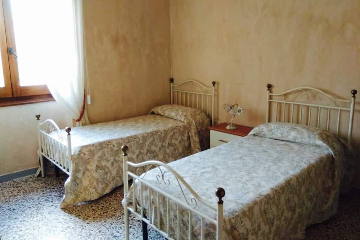 Deux lits simples avec des cadres en métal, dans une pièce aux murs unis et dotée d’une fenêtre.