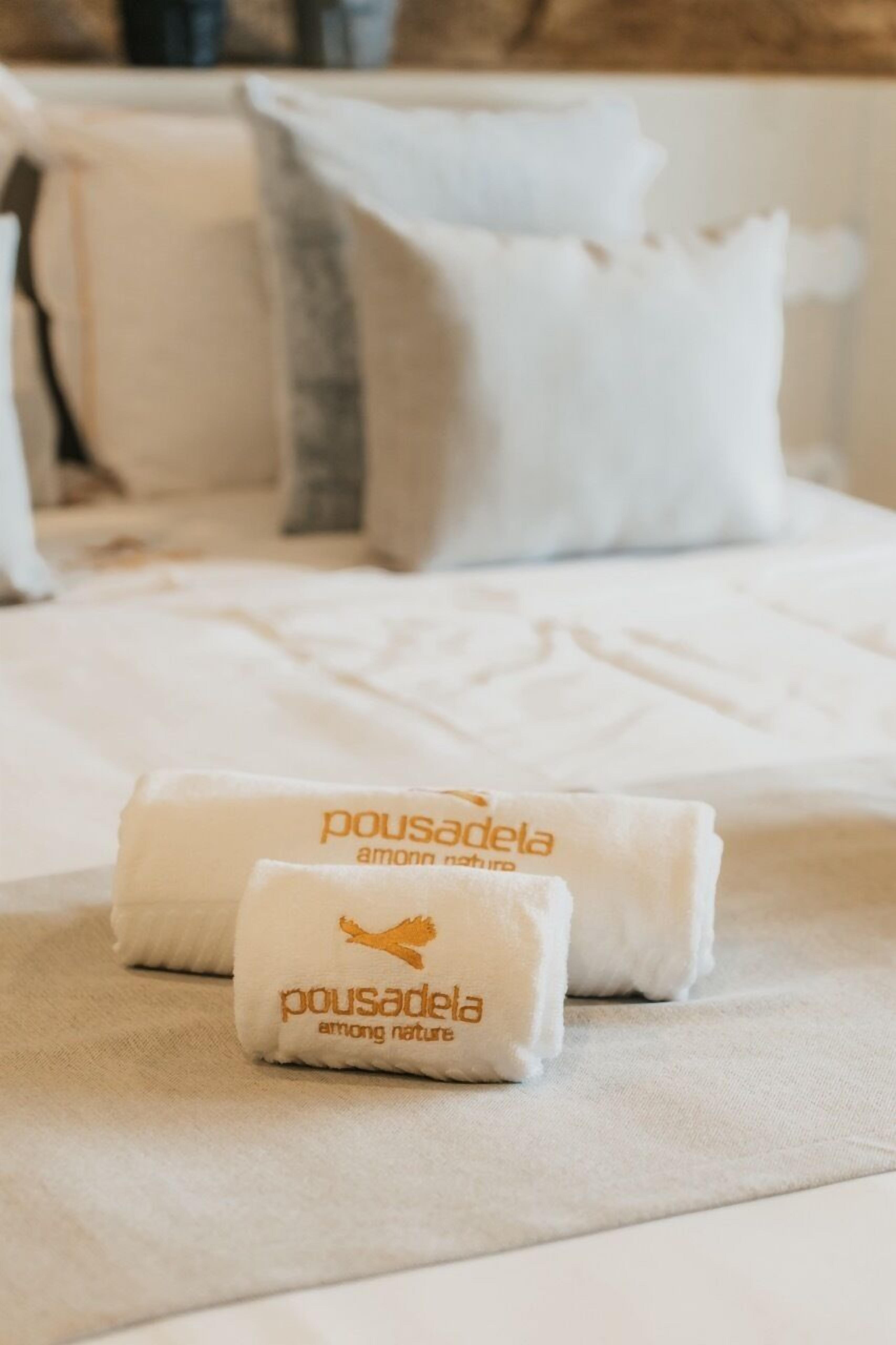 A neatly made bed with white bedding and two folded towels with the text "pousadela among nature".