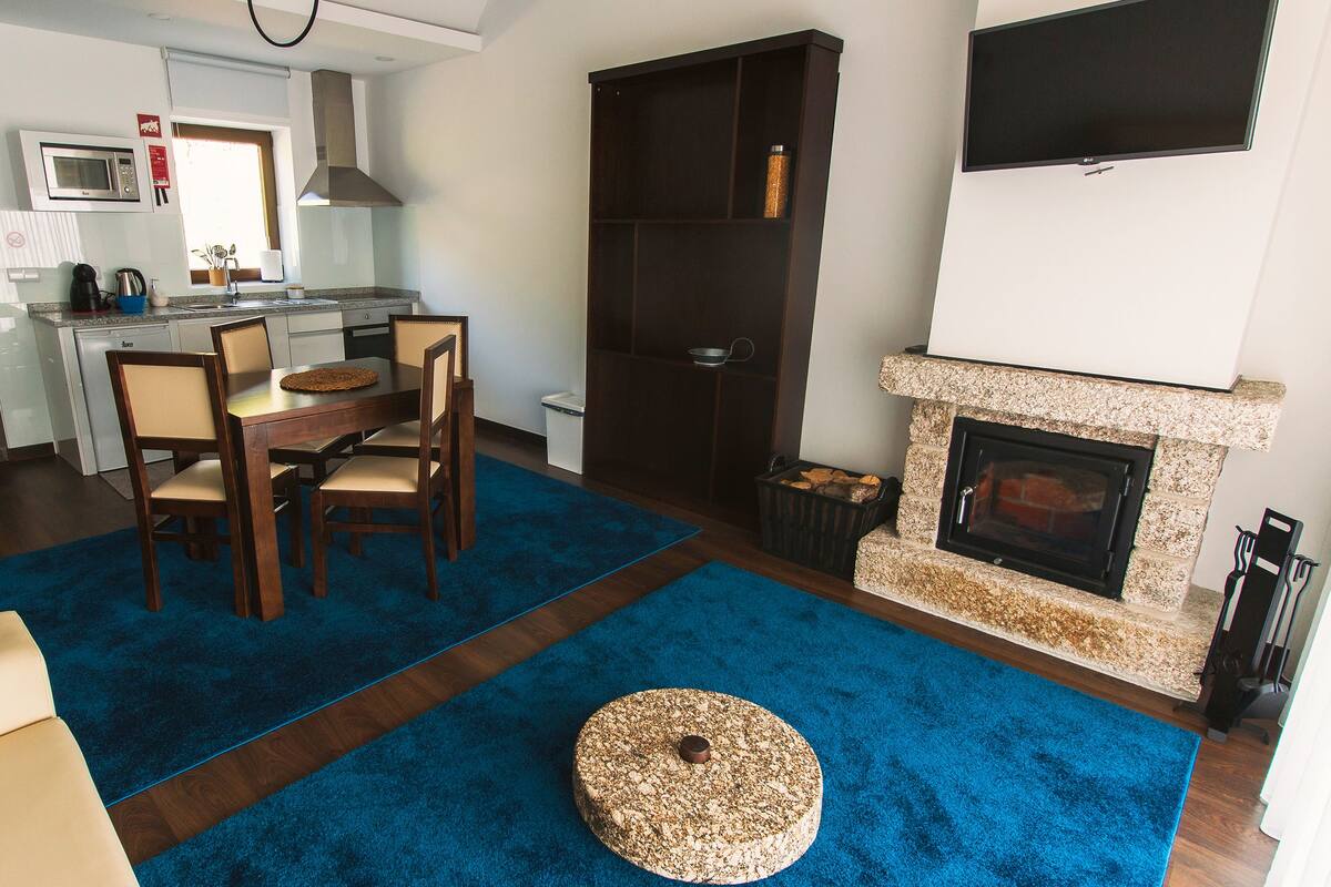 A modern living room with a blue carpet, a round ottoman, a fireplace, and a dining area with chairs and a table.