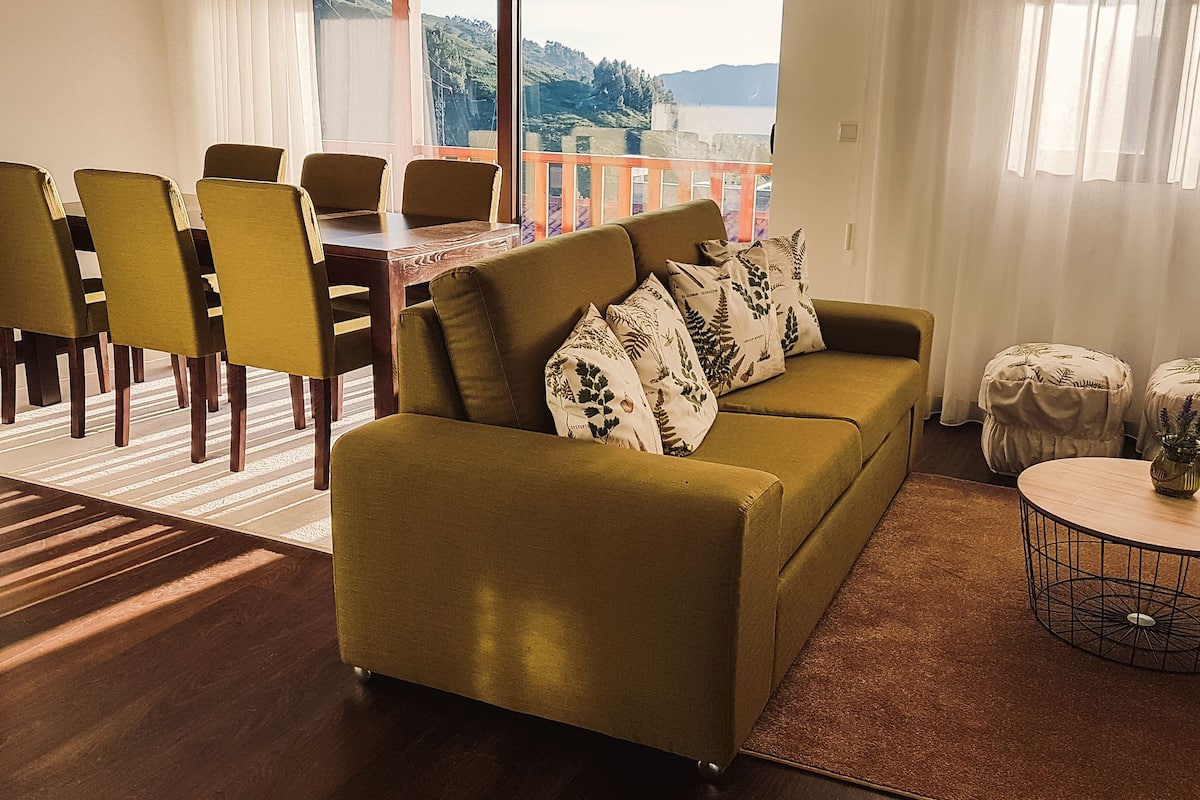 A living room with a sofa, chairs, a dining table, and a view of the sea.