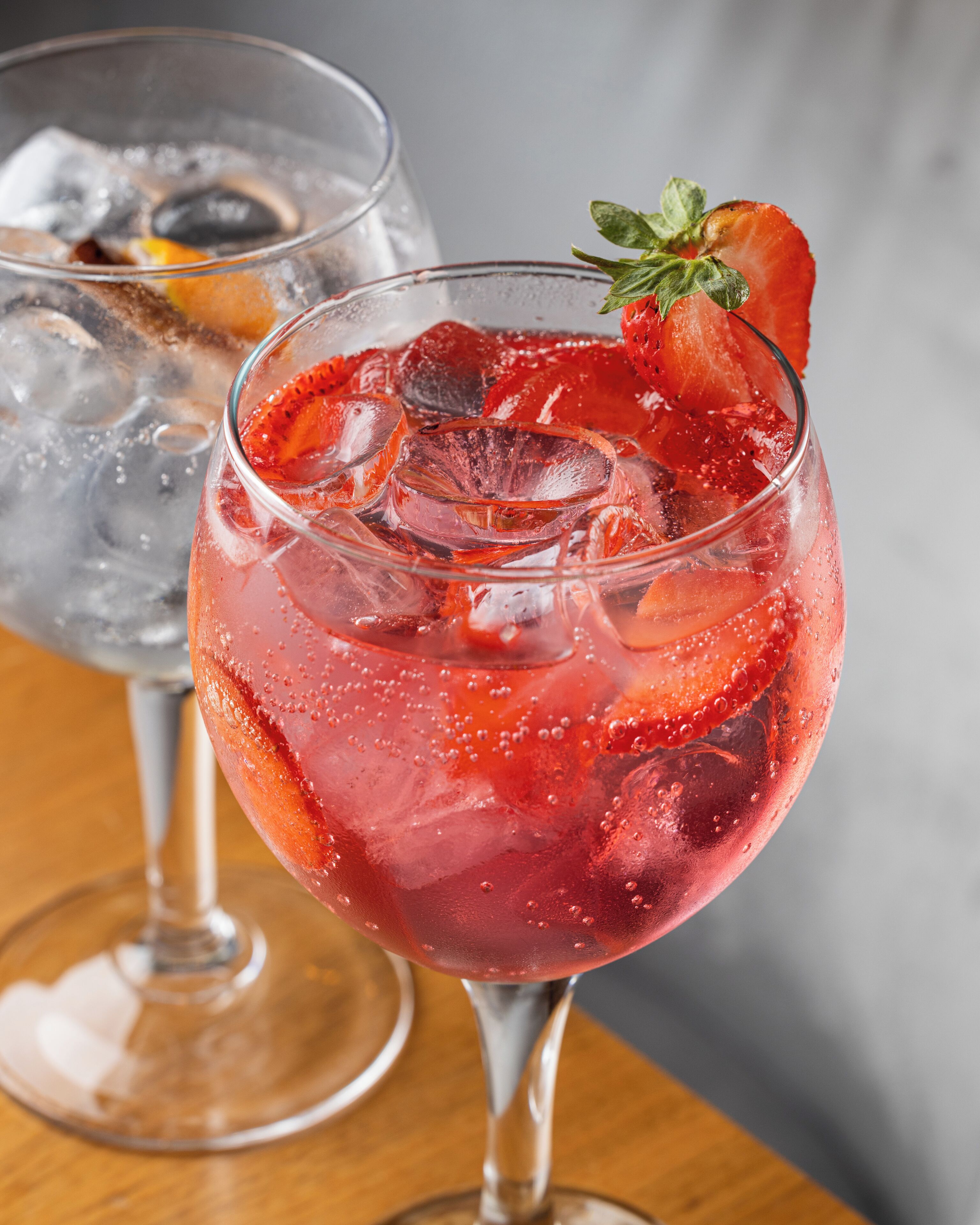 Two glasses of red beverage with ice cubes and a strawberry garnish, placed on a wooden surface.