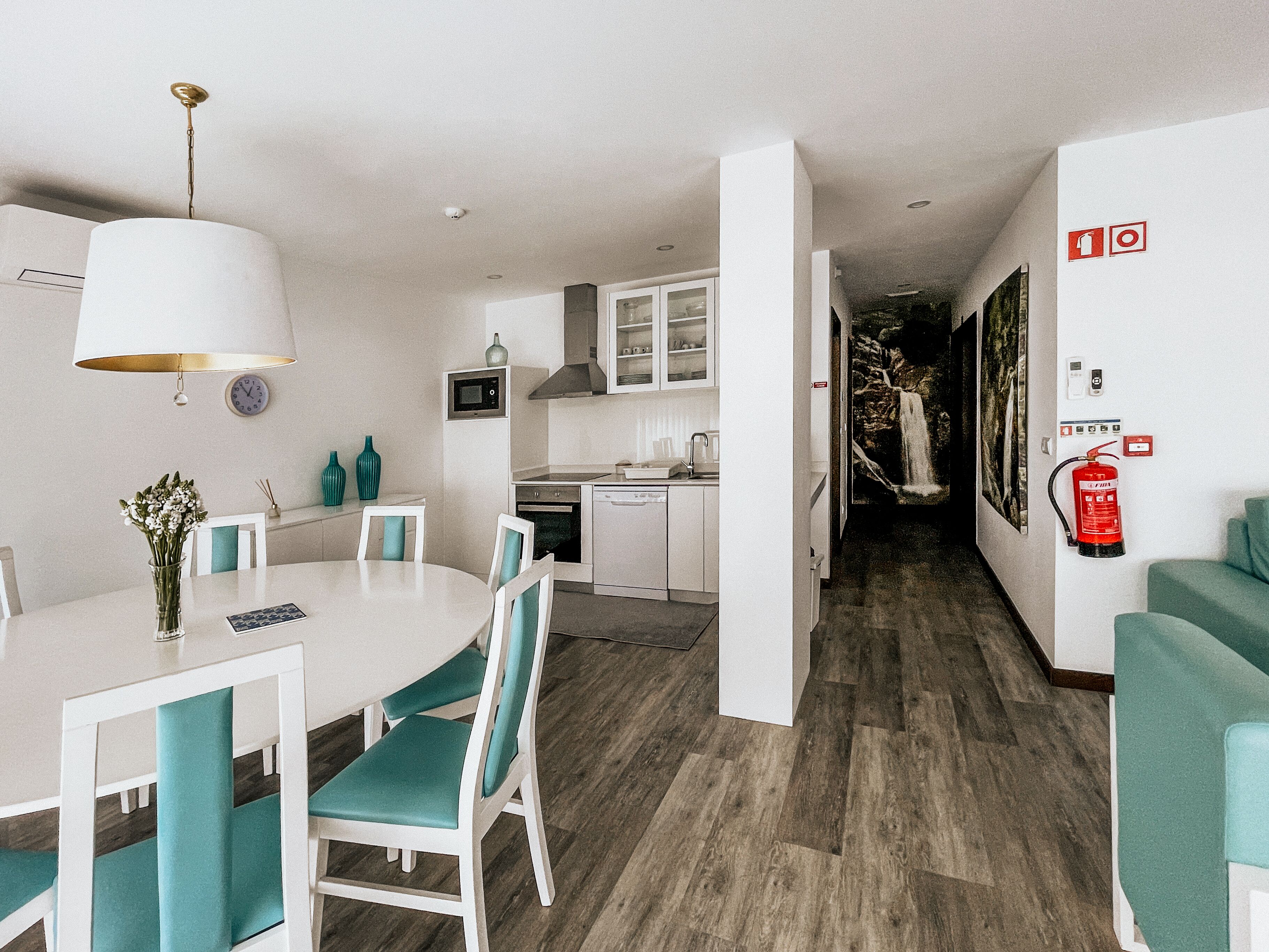 A modern kitchen with white cabinets, a microwave, and a sink. There is a dining area with a white table and teal chairs.