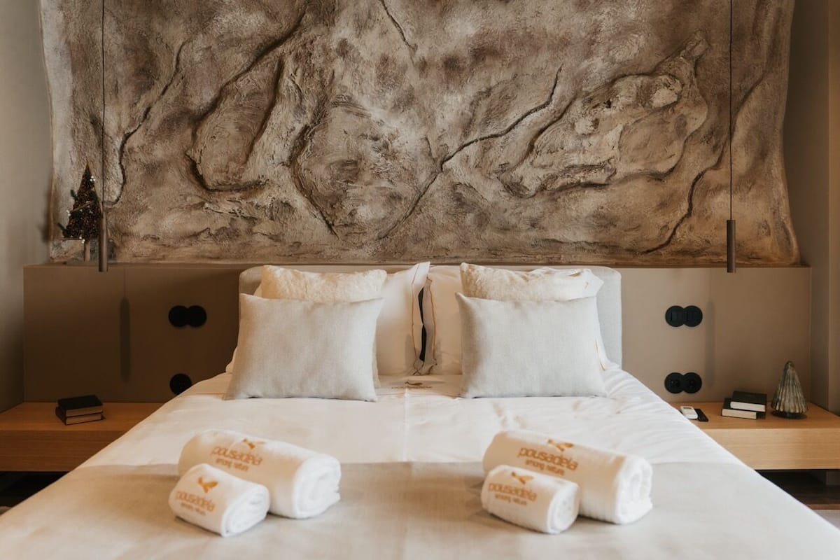 A hotel room with a large, textured wall art above the bed, a neatly made bed with white linens and two pillows, and a small nightstand with a book and a decorative item.