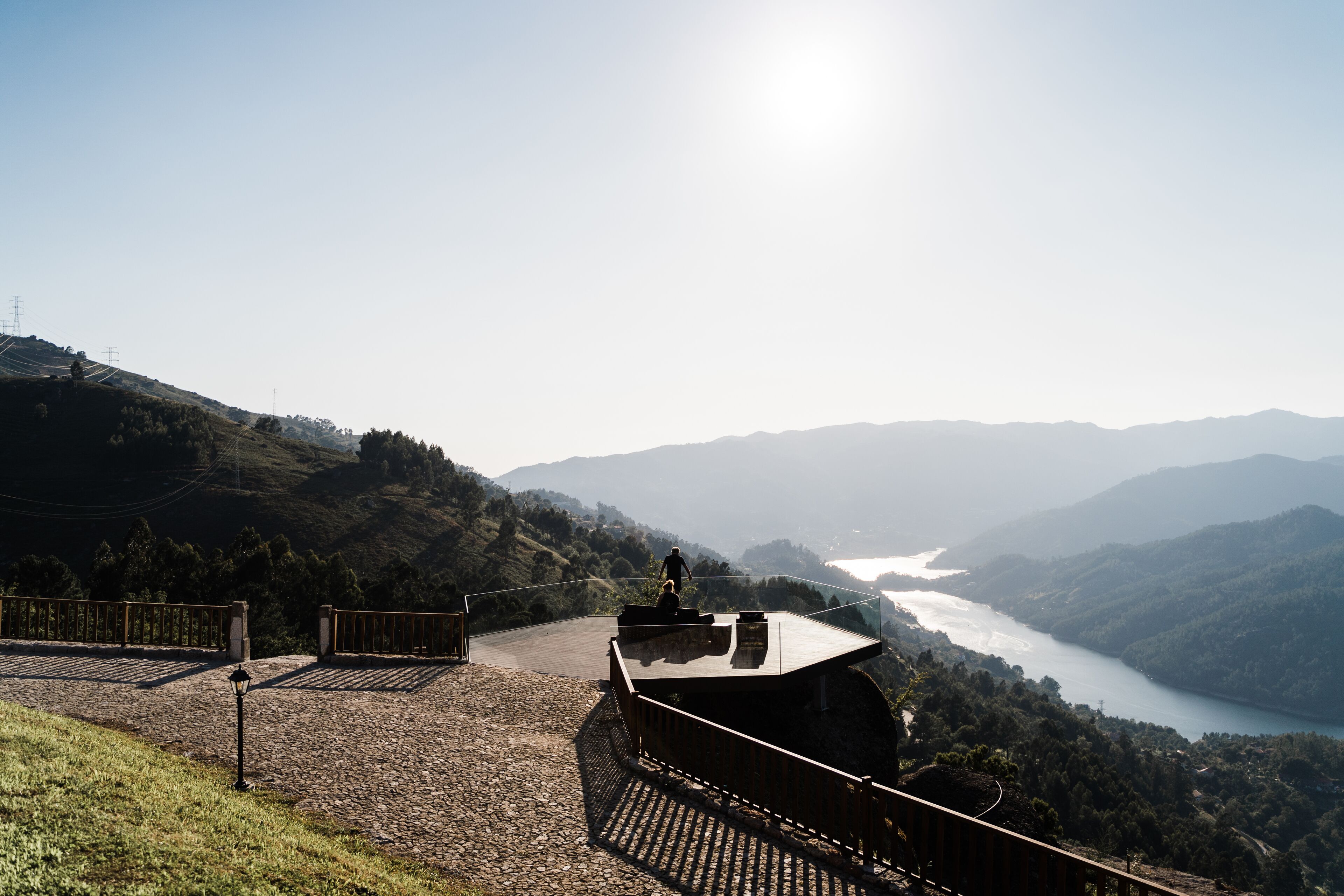 Un mirador panorámico con un sendero pavimentado, una pequeña estructura con techo de vidrio y vistas a un lago y montañas al fondo.