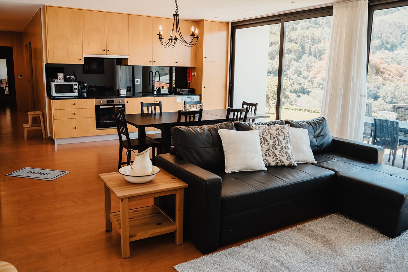A modern kitchen with wooden cabinets, a dining area with a black leather sofa, and a view of trees through large windows.