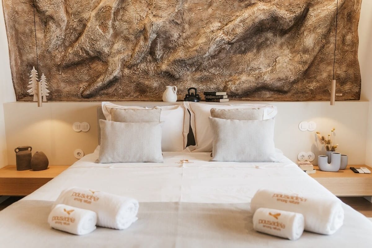 A hotel room with a large textured wall art, two beds with white linens, and a bedside table with a kettle, books, and a vase with dried flowers.