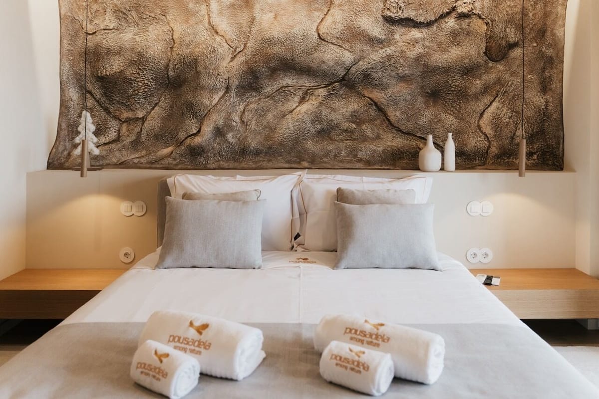 A hotel room with a large abstract painting on the wall, a bed with white linens, two pillows, and two bedside tables with white vases.