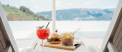 A beachside view with a table set for a meal, including a burger, chips, and a drink, overlooking a calm sea and distant mountains.