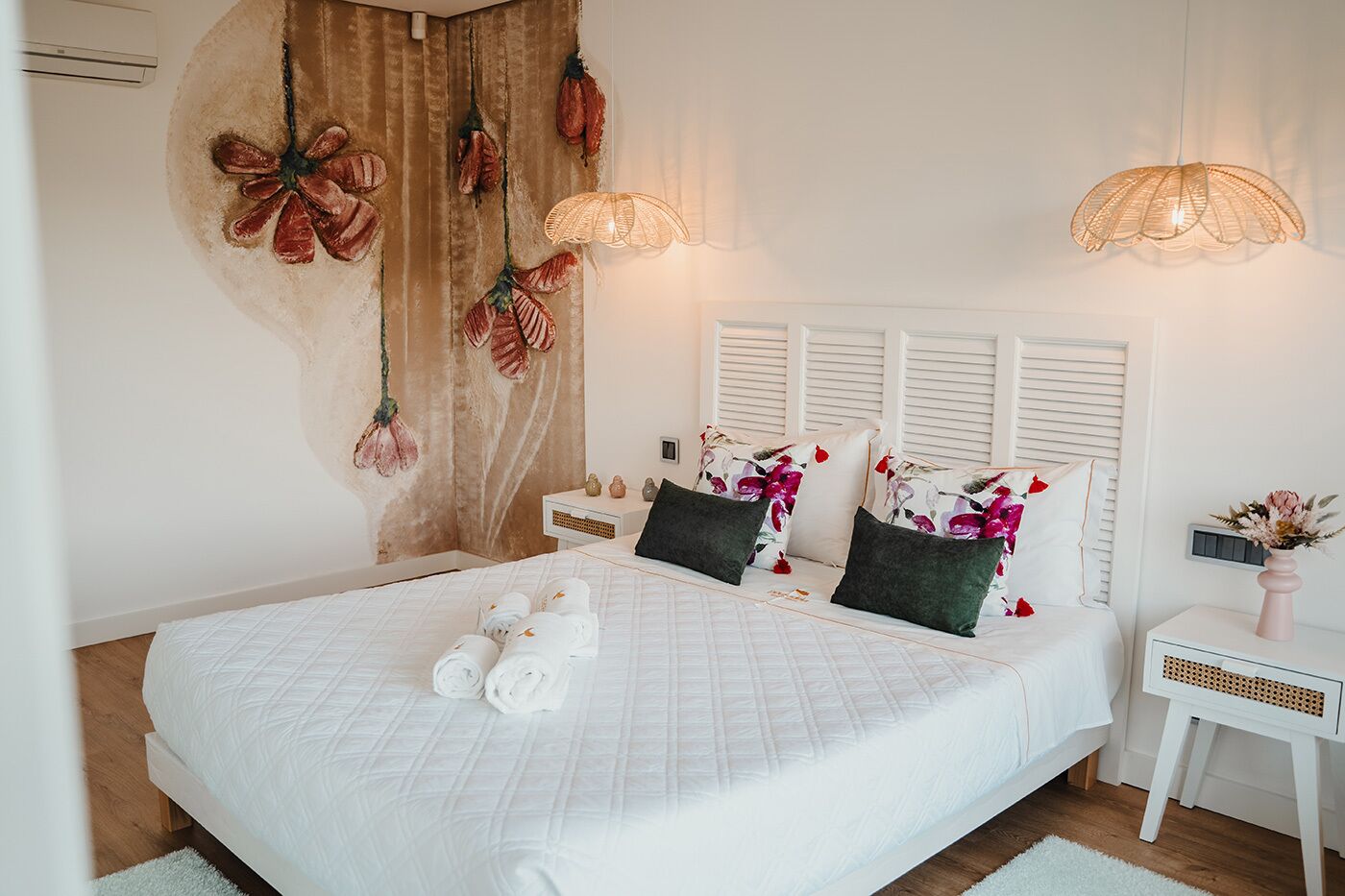A neatly made bed with rolled towels, a bedside table with a vase, and a wall with decorative elements.