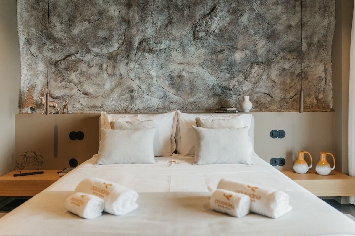 A hotel room with a large textured wall, a bed with white linens, two pillows, and two towels on the bed.