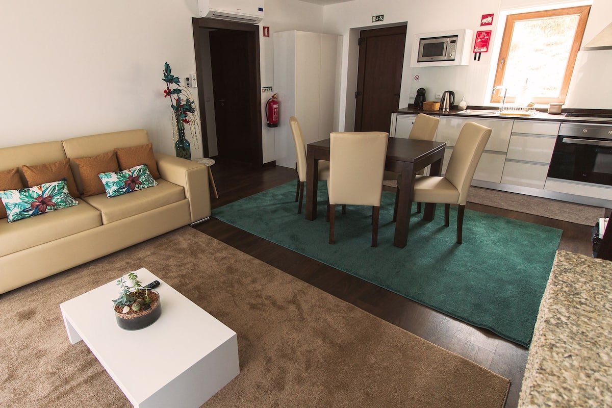 A living room with a sofa, coffee table, and dining area with chairs. There is a kitchen area with a microwave and cabinets.