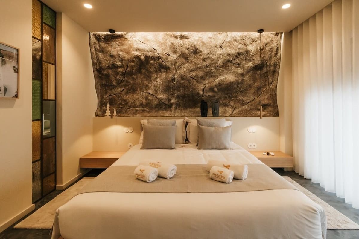 A modern hotel room with a large bed, two bedside tables, a wall-mounted artwork, and a view of the city.