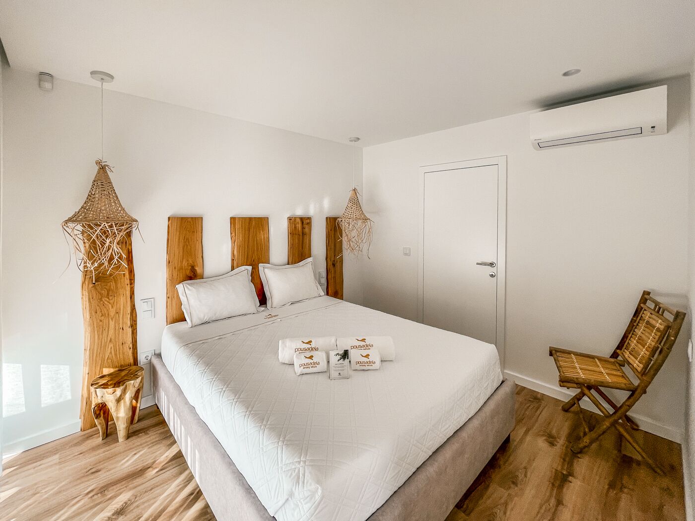 A bedroom with a large bed, wooden headboard, a wooden side table, a woven hanging light, a wooden chair, and a white door.