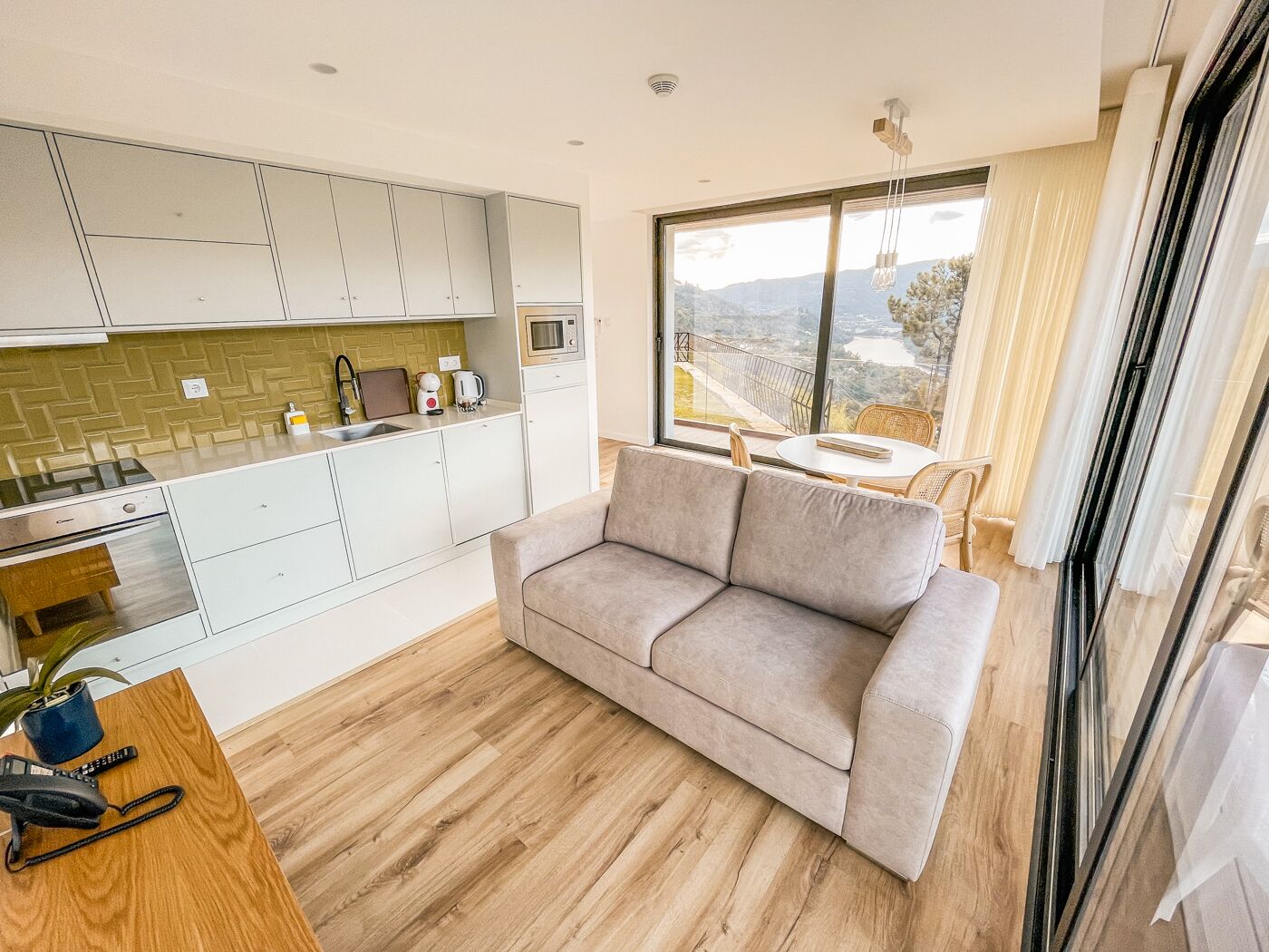 Una cucina moderna con mobili bianchi, un forno a microonde e un lavello. C'è un divano beige e un tavolo di legno con un telefono e una pianta. Un'ampia finestra offre vista sull'esterno.