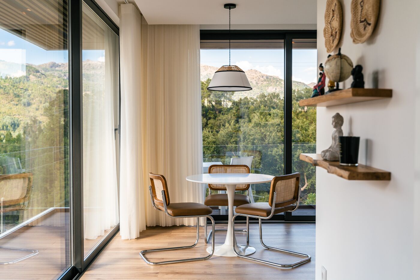 A modern dining area with a round table and chairs, large windows with curtains, and a view of the outdoors.
