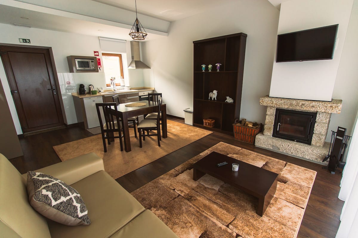 A modern living room with a sofa, coffee table, and fireplace. There is a dining area with chairs and a table, and a kitchen in the background.
