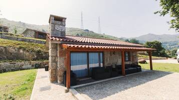 A stone building with a tiled roof, a covered patio with seating, and a view of hills and power lines in the background.