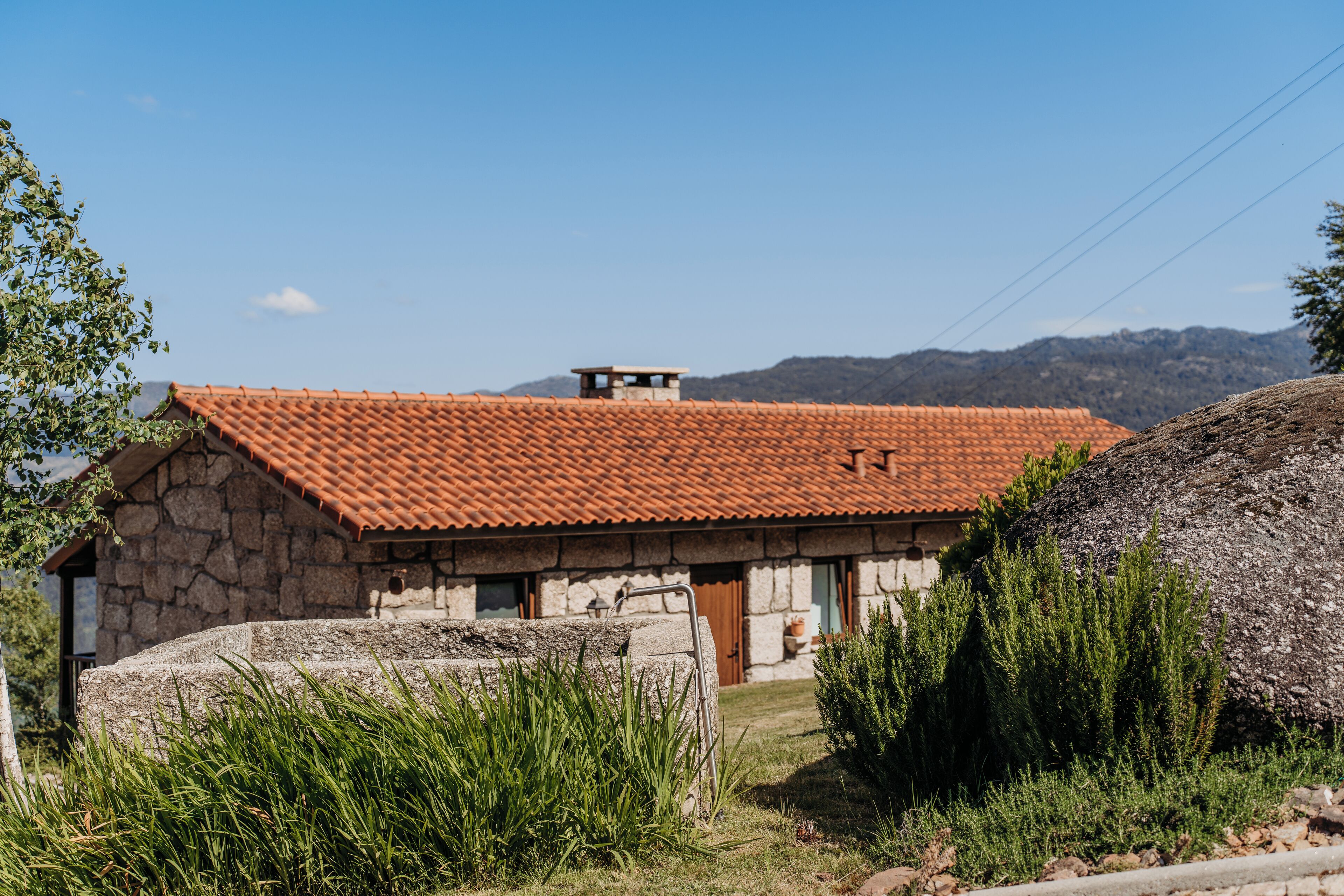 Um edifício de pedra com telhado em tijolos vermelhos, cercado por vegetação e ao fundo montanhas.