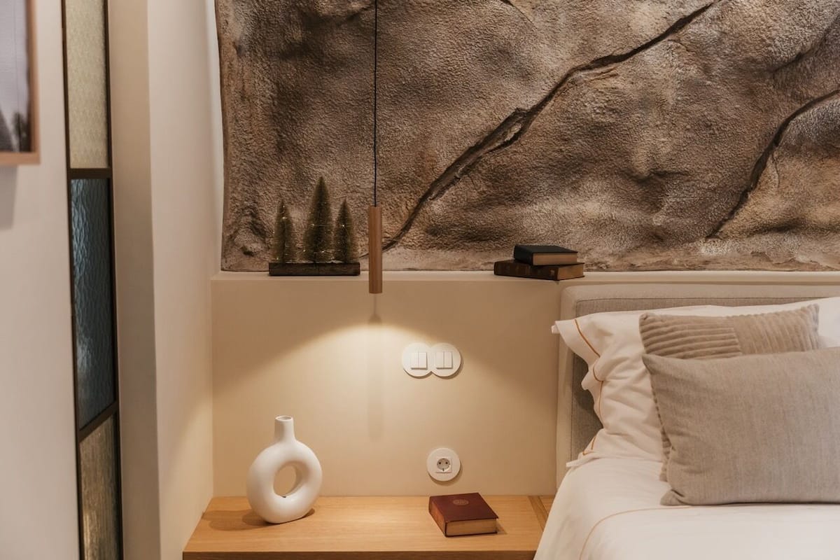 A neatly arranged bedroom with a large textured wall art, a bedside table with a vase and books, and a bed with pillows.