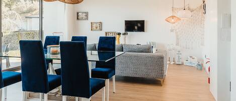A modern dining area with a glass table and blue chairs, a gray sofa, and a hanging light fixture.