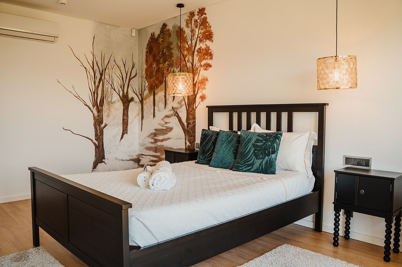 A modern bedroom with a dark wooden bed, white bedding, and green leaf-patterned pillows. There are two pendant lights and a wall-mounted air conditioner.