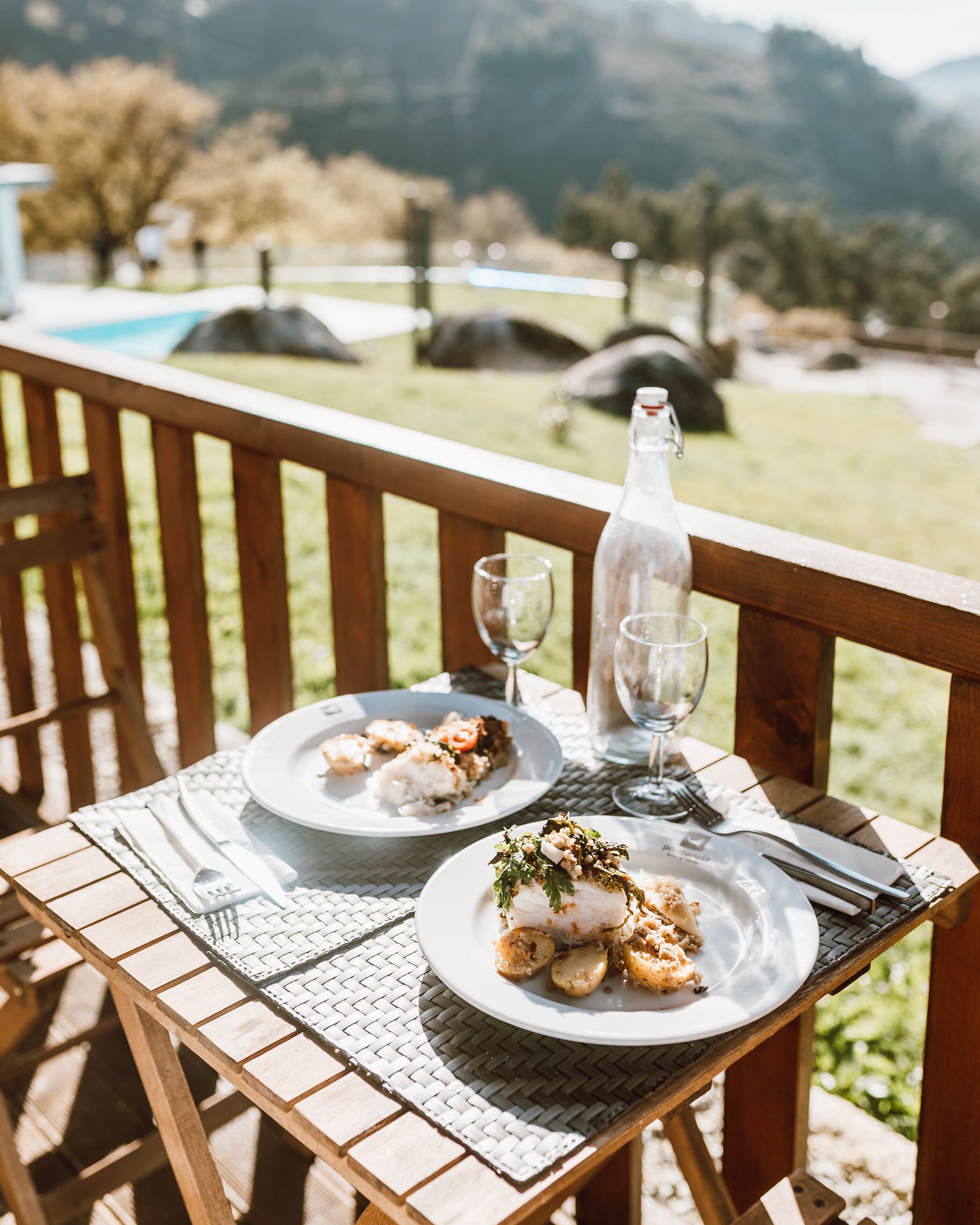 Eine Holzterrasse mit Tisch für zwei Personen, gedeckt mit Speisen, einer Flasche und Weingläsern.