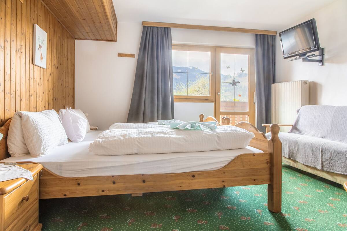 Uma cama de madeira com roupa de cama branca, uma mesinha de cabeceira de madeira, uma cadeira, uma televisão na parede e uma janela com cortinas.