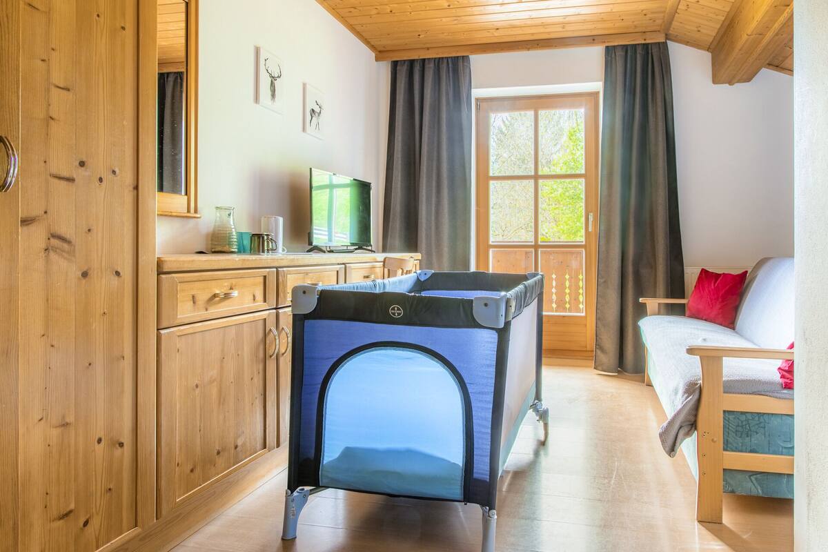Quarto aconchegante com decoração em madeira, cozinha compacta, televisão, janela com cortinas e cama com travesseiro vermelho.
