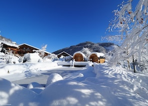 Una cabaña nevada con cabañas de madera, cielo azul despejado y montañas al fondo.