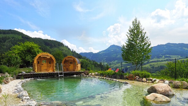 A clear pool with wooden cabins, surrounded by rocks and greenery, with mountains in the background. - Gasthof Pesbichl (Goldegg)