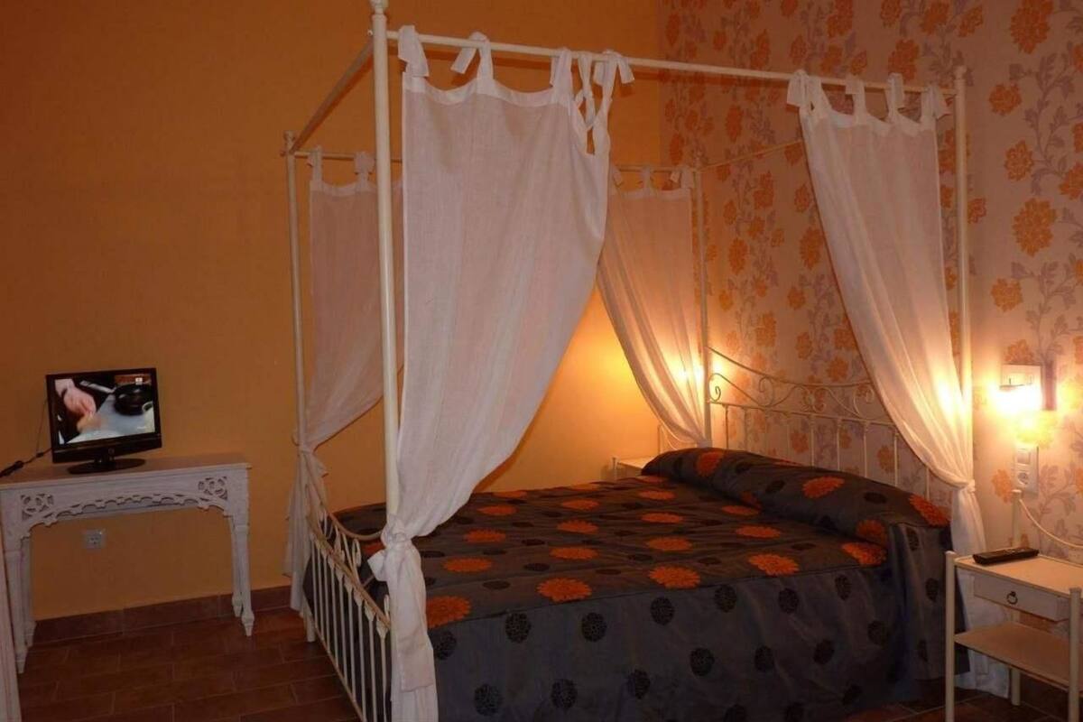 Een hemelbed met witte gordijnen, een televisie op een bijzettafeltje en een nachtkastje met een lamp.