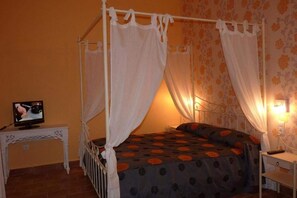 Una cama con dosel adornada con cortinas blancas, un televisor sobre una mesita y una mesilla de noche con lámpara.