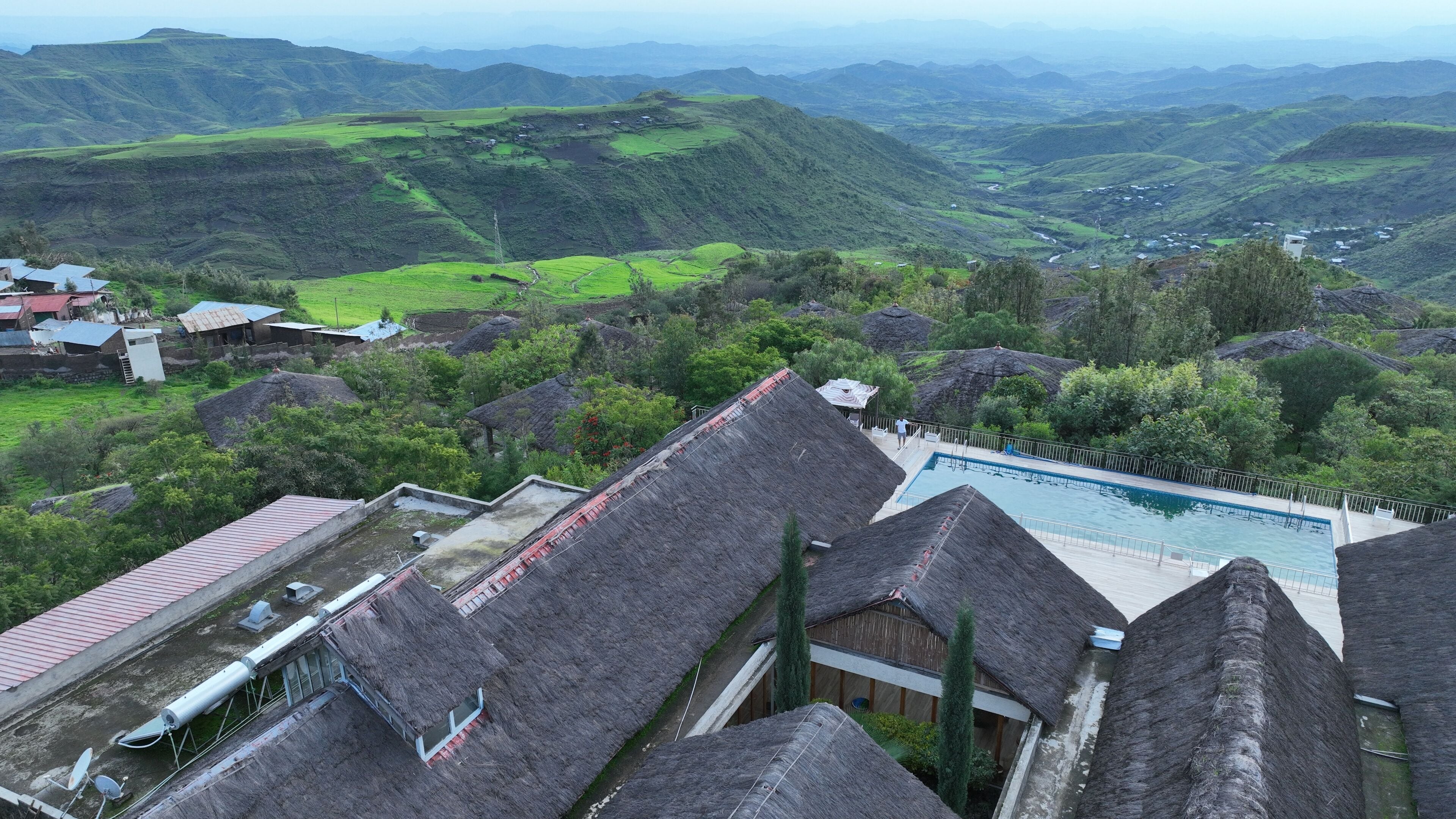 A traditional village with thatched-roof houses, a swimming pool, and lush green hills in the background.