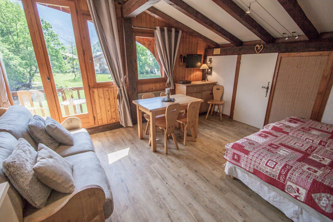 Une pièce chaleureuse avec un plafond en bois, un lit recouvert d’une couverture à motifs, une table à manger avec des chaises, et une vue sur la verdure à l’extérieur.