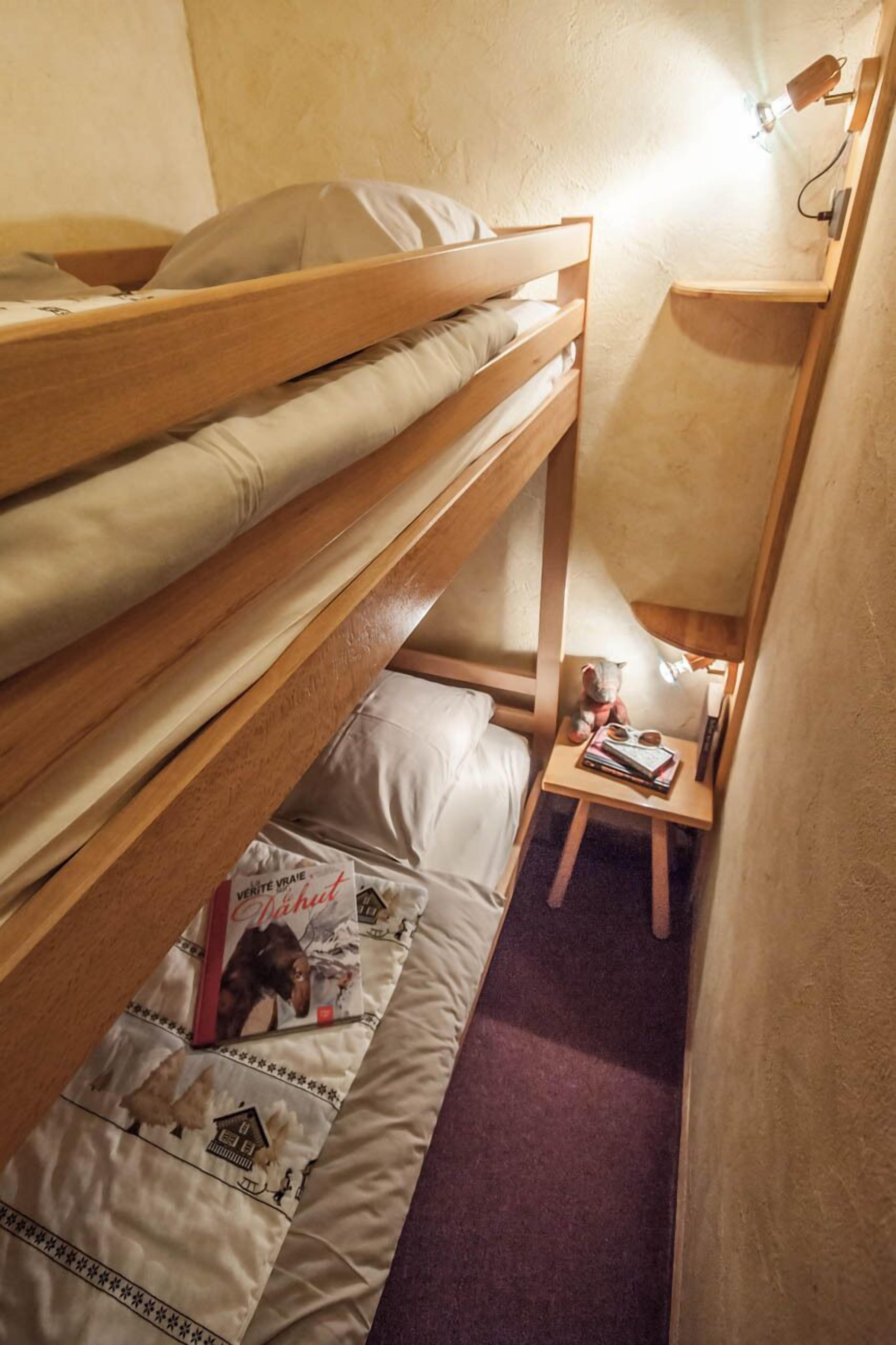 Une petite pièce étroite, avec un lit superposé, une petite table sur laquelle est posé un ours en peluche, et une lampe de lecture fixée au mur.