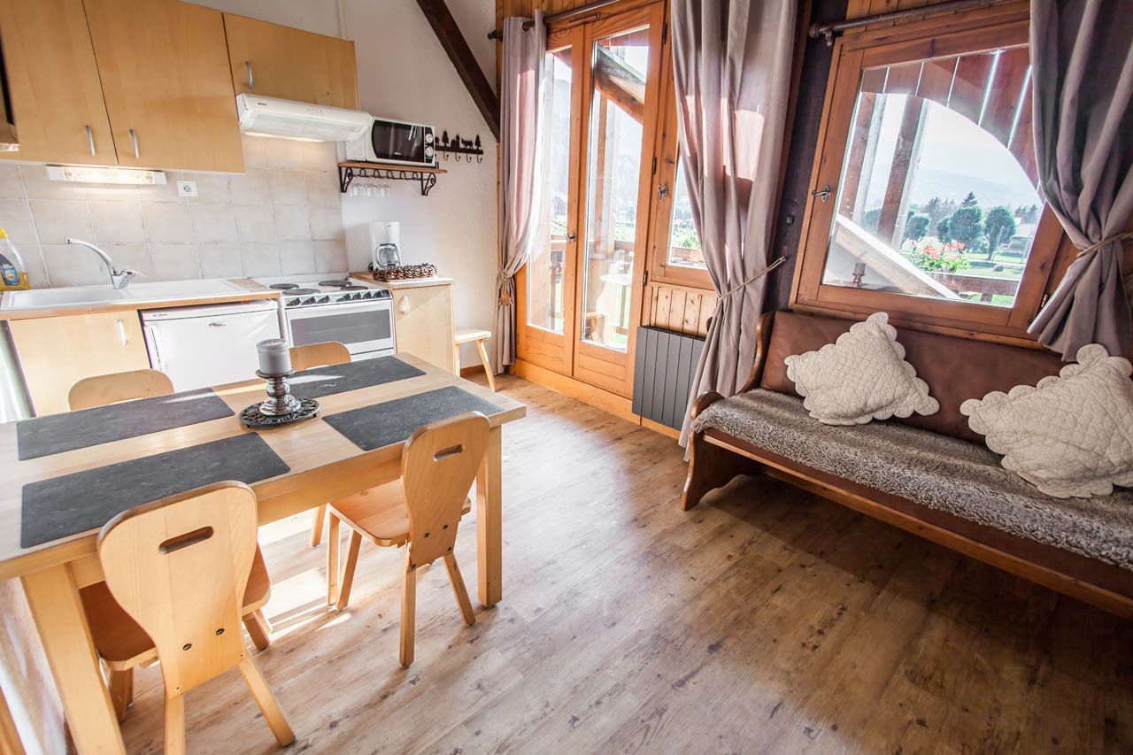 A cozy kitchen with wooden cabinets, a dining table with chairs, a sofa with cushions, and a view through a window.