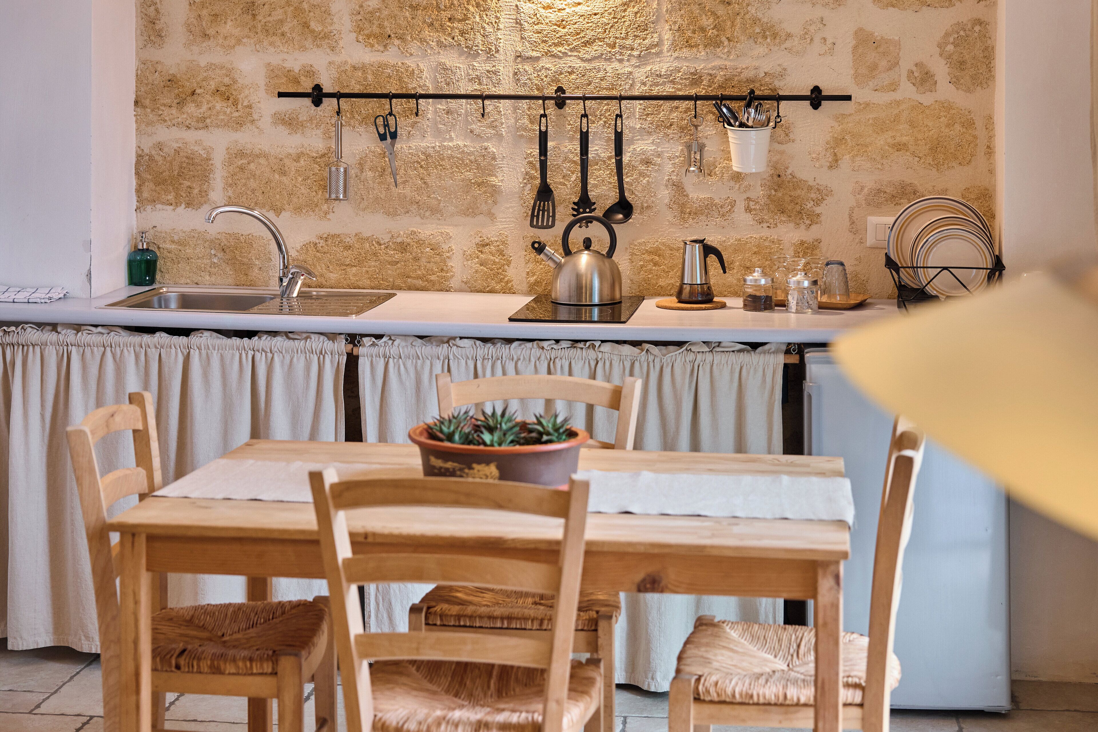 A kitchen with a stone wall, a hanging utensil rack, a kettle, and a dining table with chairs.