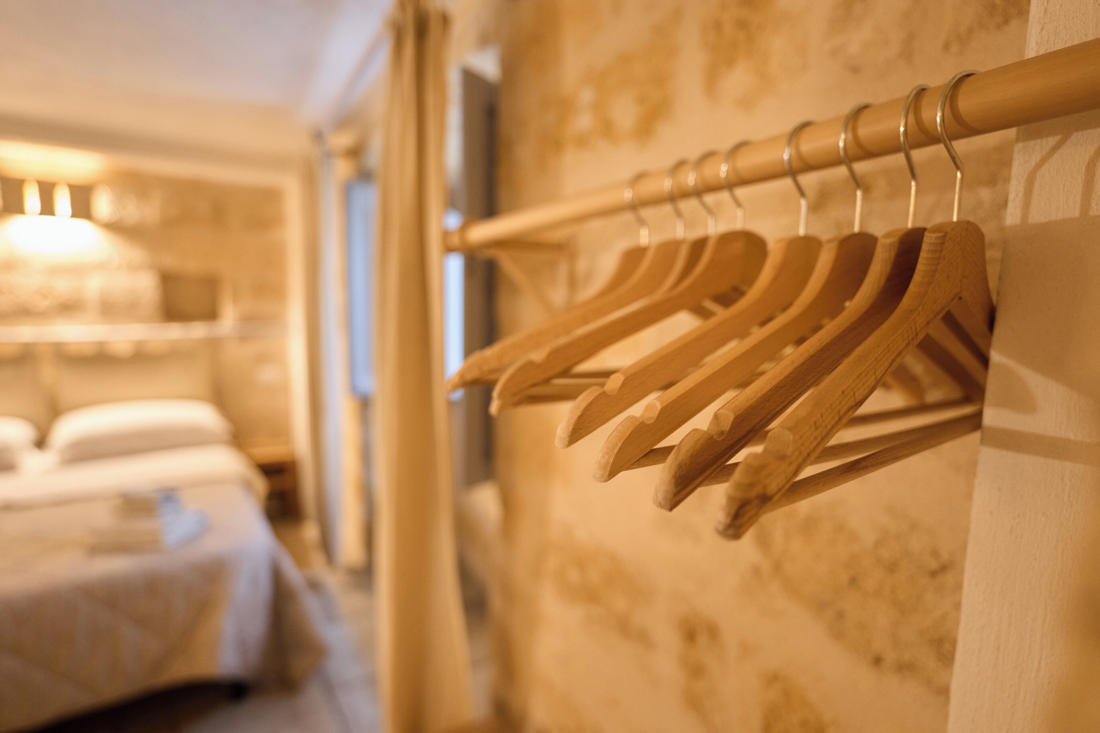 Habitación de hotel con un perchero de madera en la pared, una cama con colcha blanca y una lámpara al fondo.