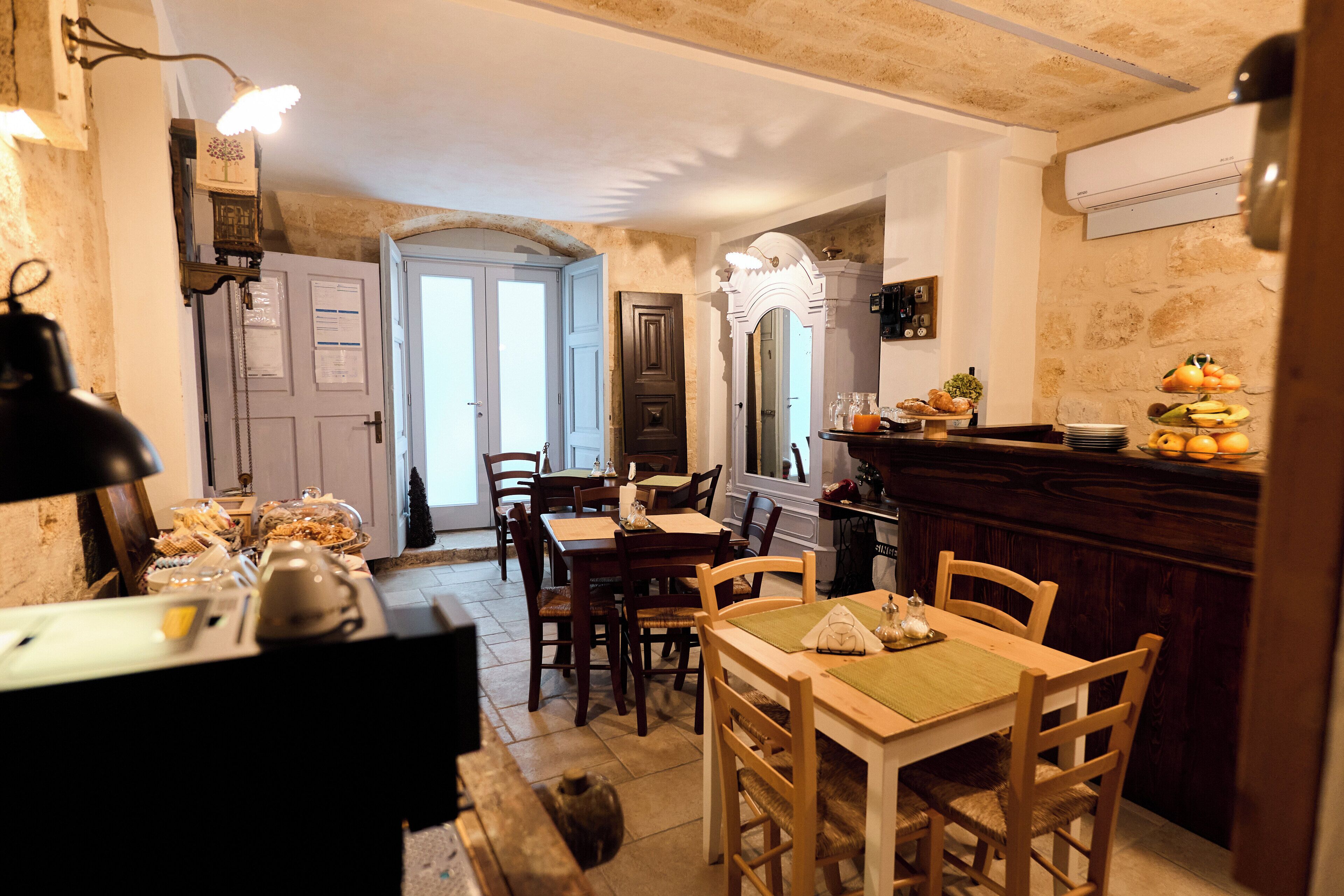 Un área de comedor con mesas y sillas de madera, una barra con fruta y pasteles, y un espejo sobre la barra.