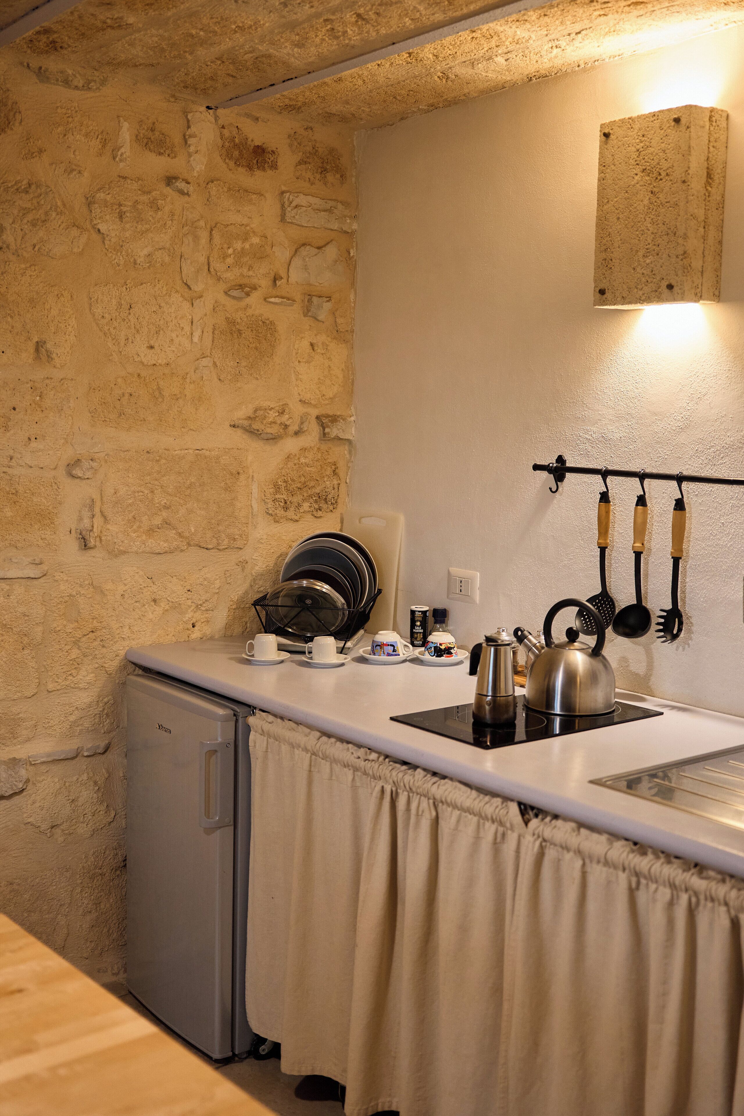 A compact kitchen with a stone wall, a small refrigerator, a kettle, and a utensil rack.