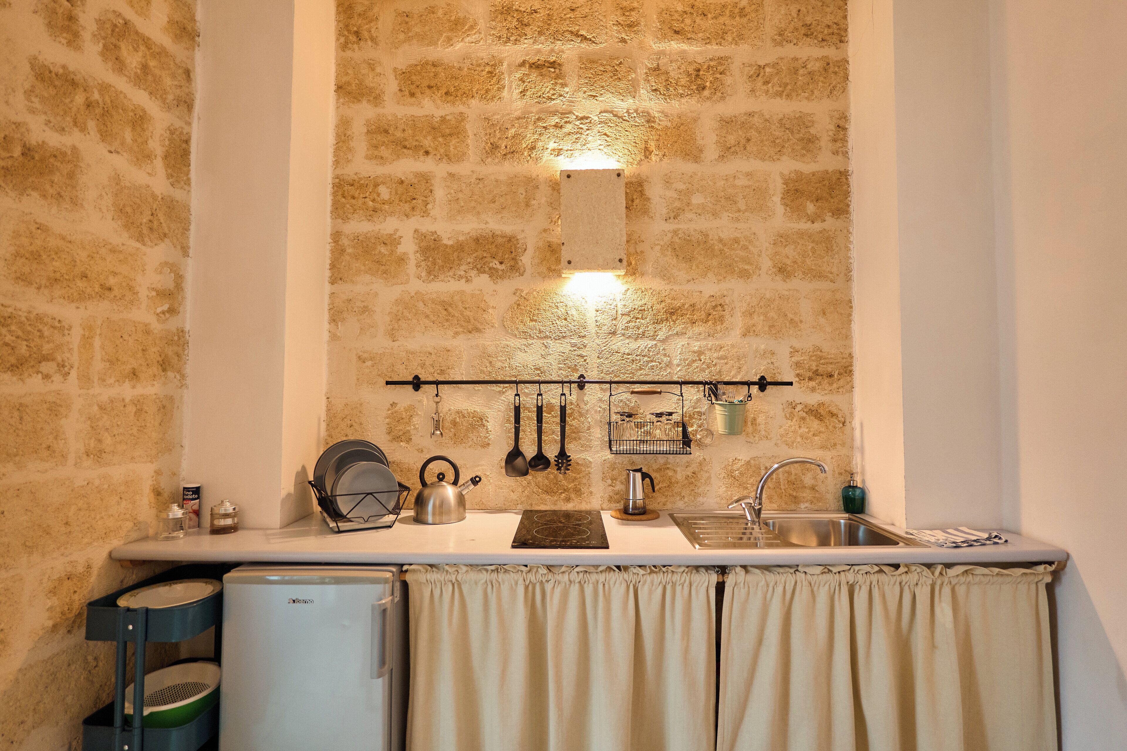 A kitchen with a stone wall, a sink, a kettle, and a small refrigerator.