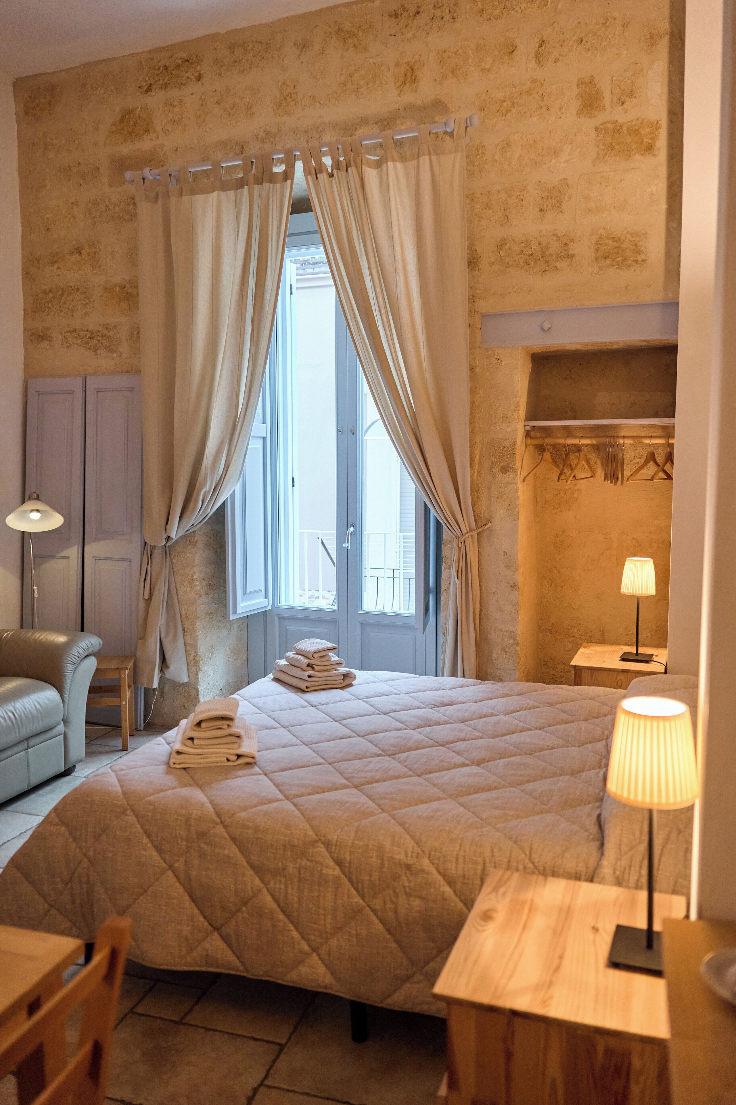 A bedroom with a large bed, a bedside table, a lamp, a chair, a small table, and a window with curtains.