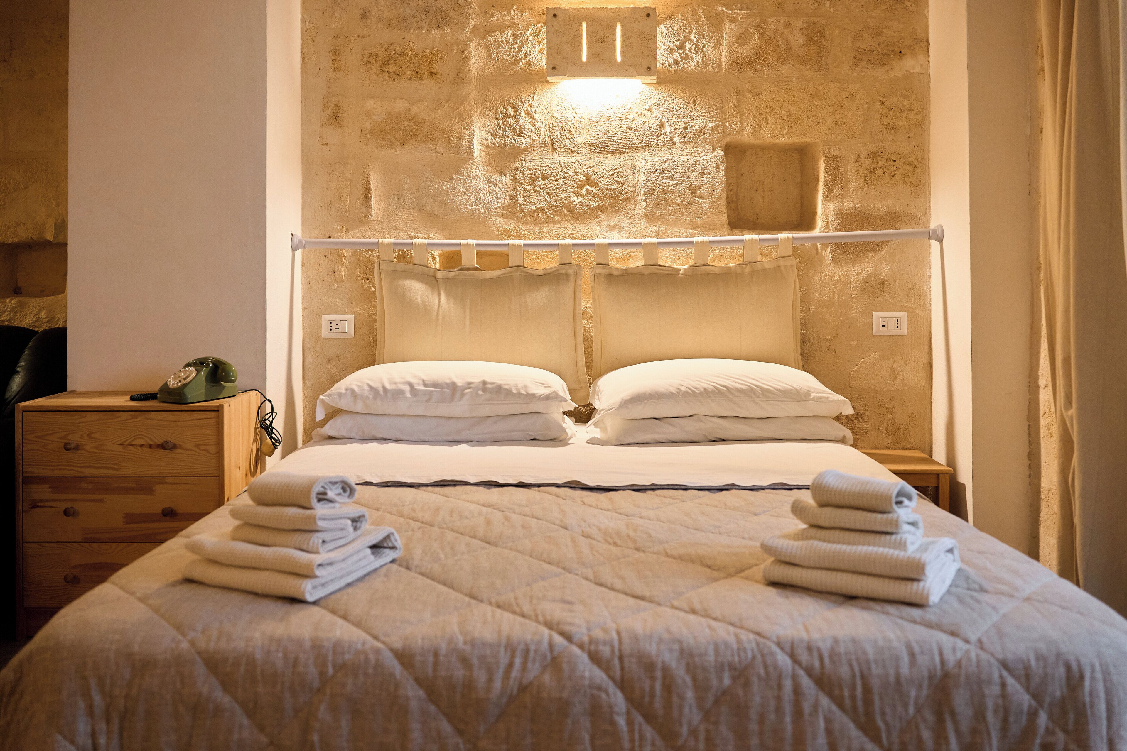 A neatly made bed with folded towels, a wooden nightstand with a lamp, and a stone wall with a light fixture.