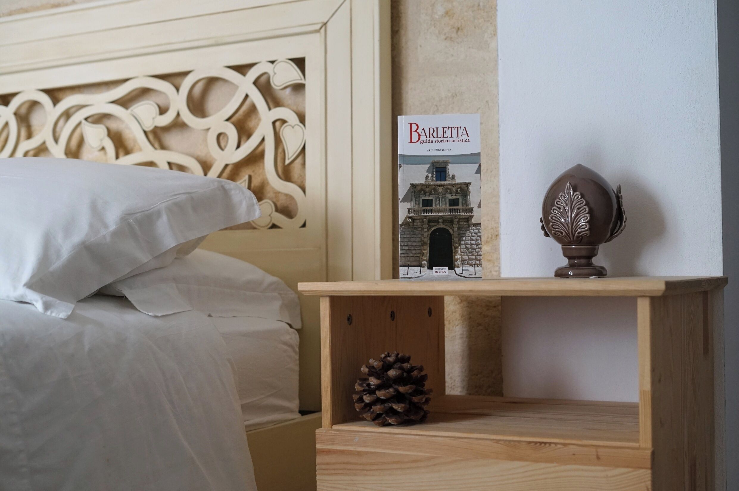 Ein ordentlich bezogenes Bett mit weißen Leinen, ein dekorativer Kopfteil, ein kleiner Nachttisch aus Holz mit einem Buch mit dem Titel 'Barletta' und einer Kiefernzapfen darauf, sowie eine Wand mit einer strukturierten Oberfläche.