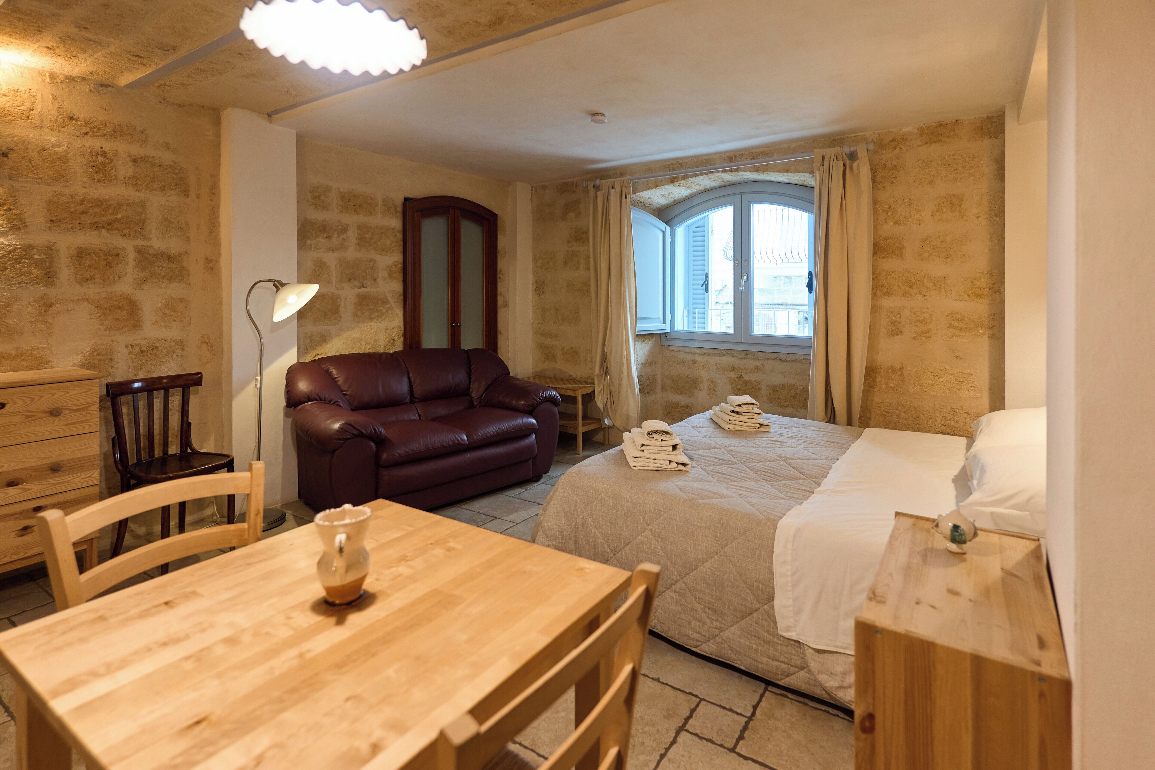 A cozy room with a wooden table, a leather sofa, a bed with a quilt, and a window with curtains.