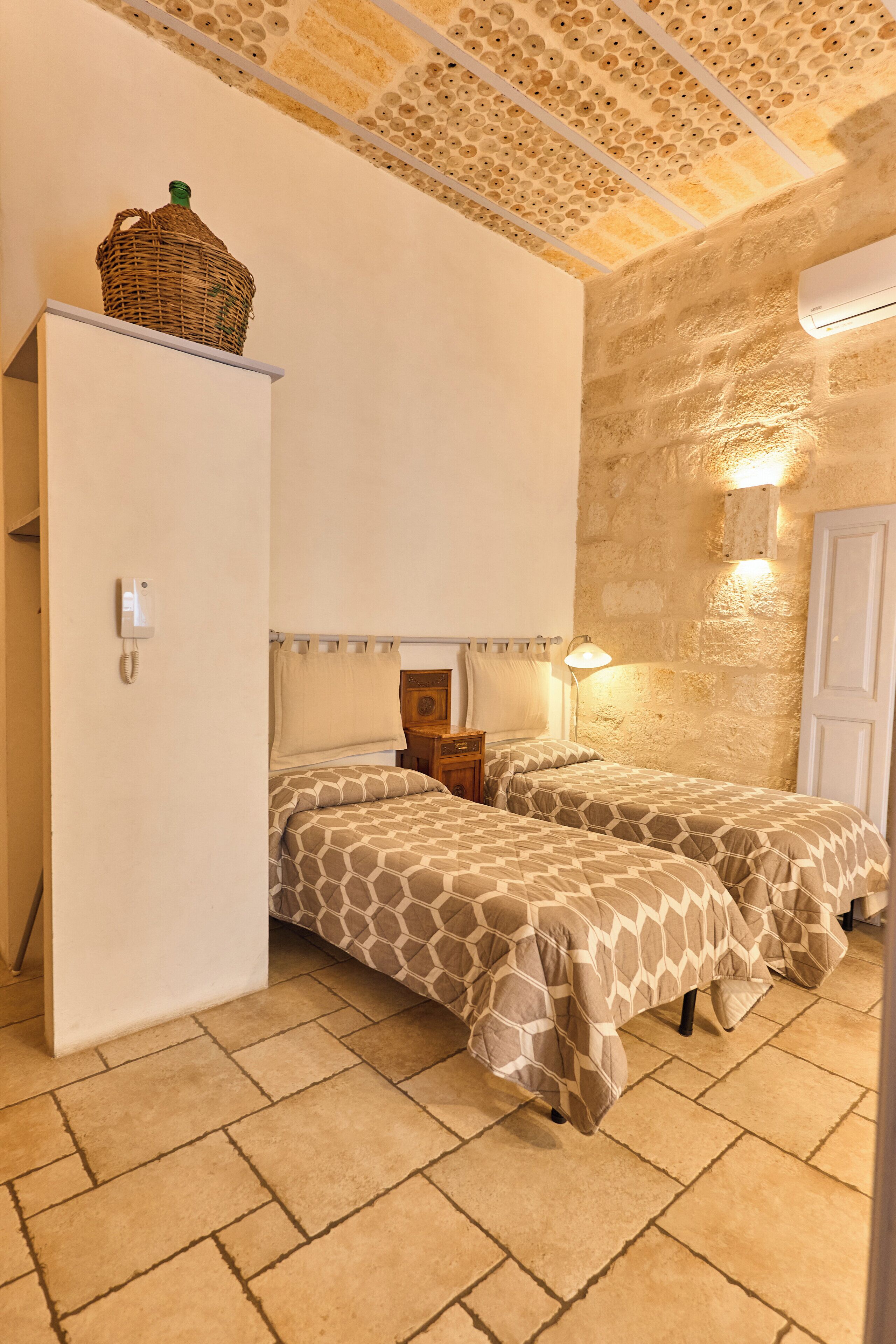 A room with two beds, a small table, a wicker basket on a cabinet, and a white door.